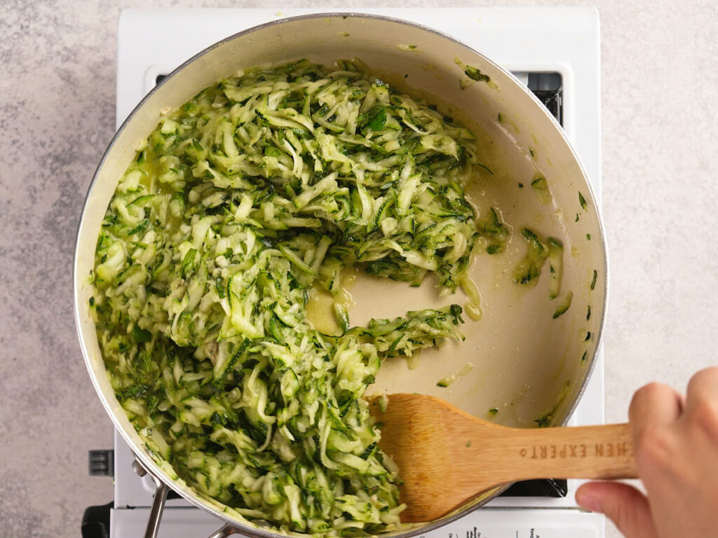 A hand stirs shredded zucchini in a large pot on a stovetop with a wooden spatula.