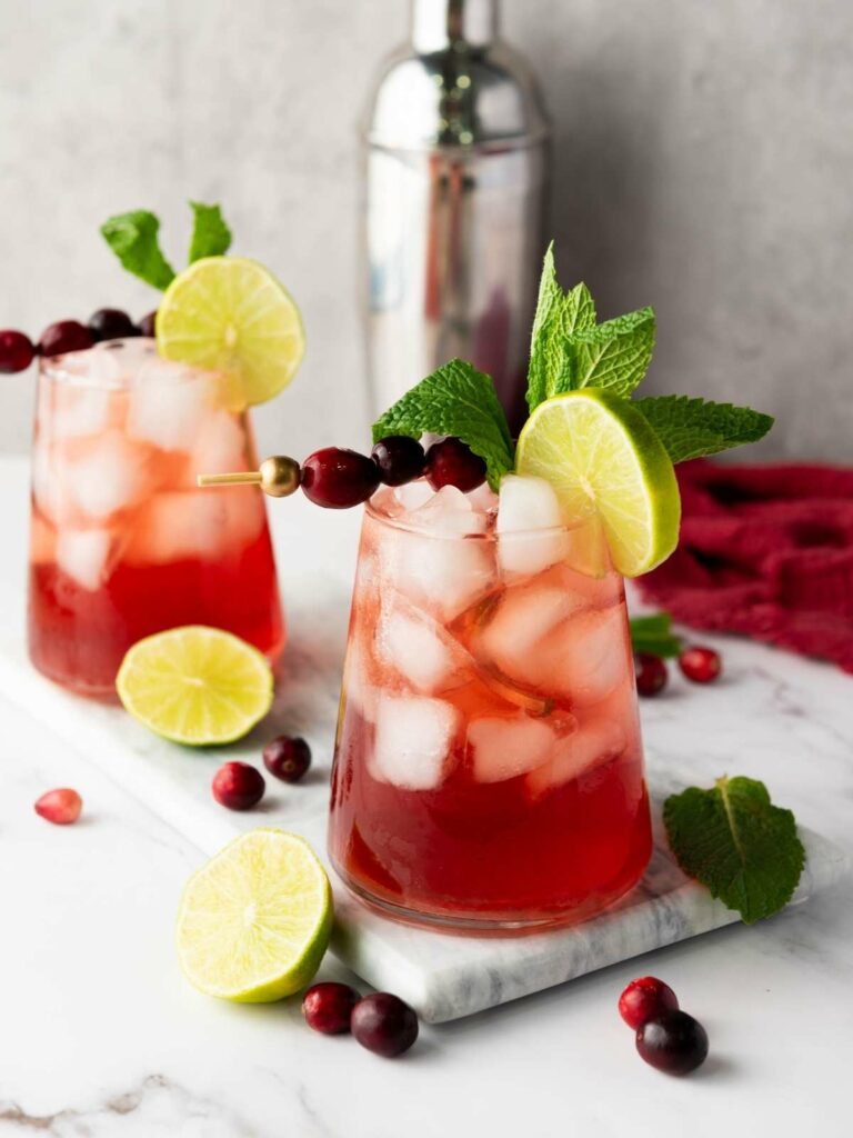 Two glasses of red iced cocktails garnished with lime slices, mint leaves, and cranberries, set on a marble surface.