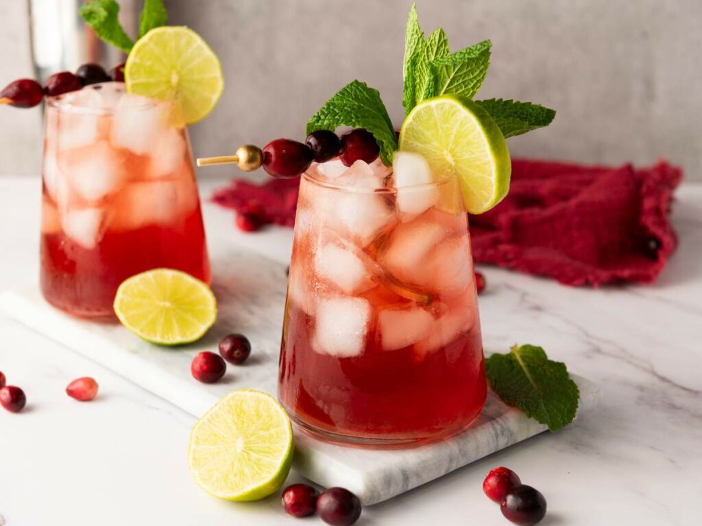 Two glasses of red iced drink garnished with lime slices, mint leaves, and cranberries, on a marble slab.