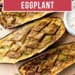 Three oven-baked eggplant halves with a crosshatch pattern, garnished with herbs, on a wooden board.