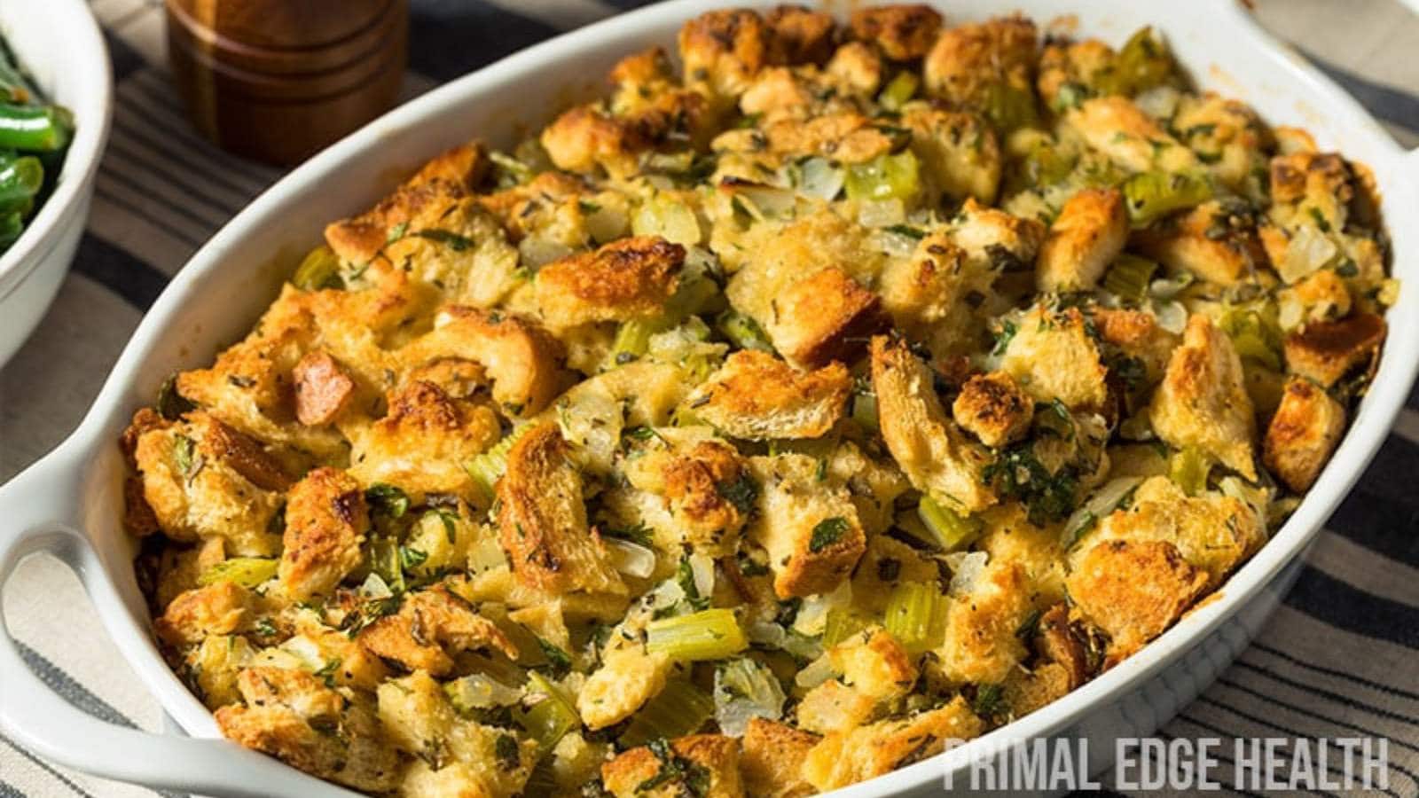 A white baking dish filled with baked bread stuffing containing chunks of bread, vegetables, and sausage.