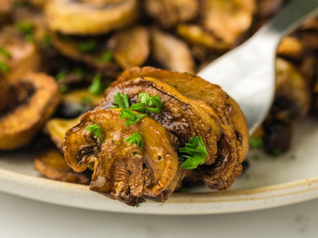 Close-up of cooked sliced mushrooms garnished with chopped parsley, served on a white plate with a spoon.