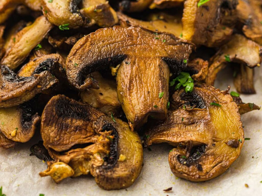 Close-up of several cooked and sliced mushrooms garnished with small bits of fresh herbs on a parchment-lined surface.
