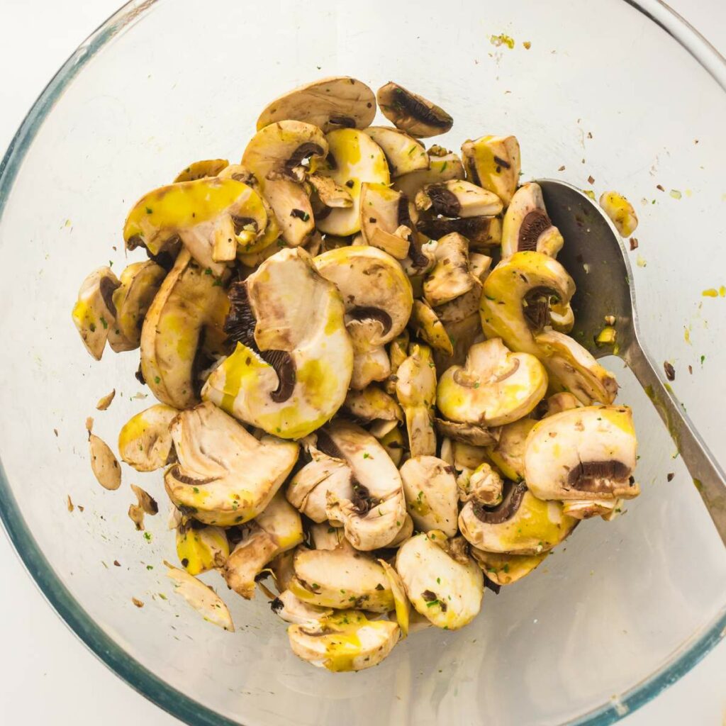 Sliced mushrooms mixed with herbs and yellow seasoning in a glass bowl with a metal spoon.