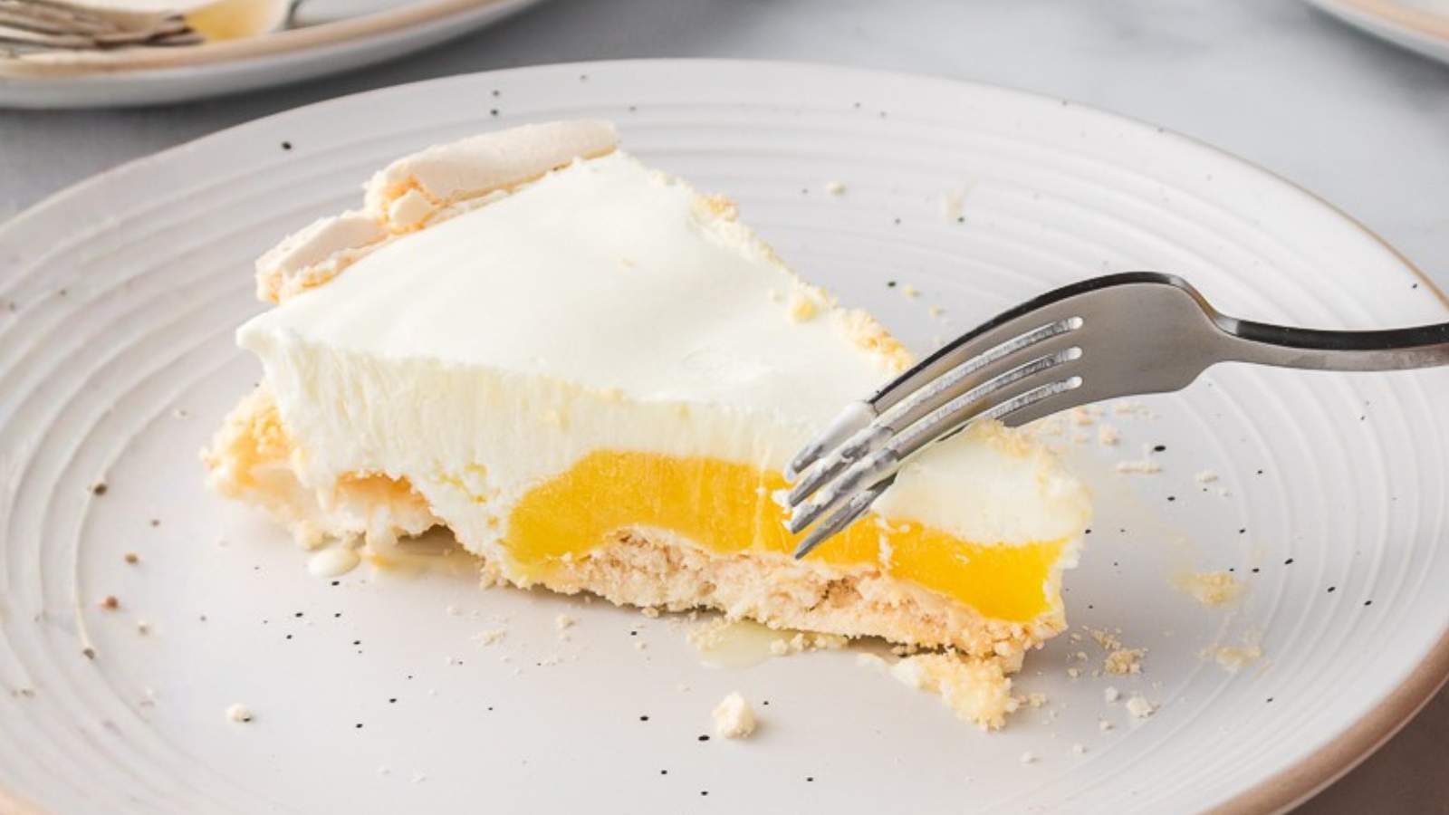 A fork cutting into a slice of angle pie with a white creamy topping and a yellow filling on a white plate.