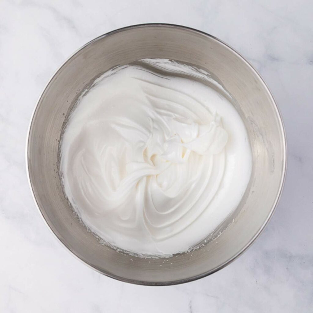A stainless steel bowl filled with stiff, glossy whipped egg whites on a white marble surface.