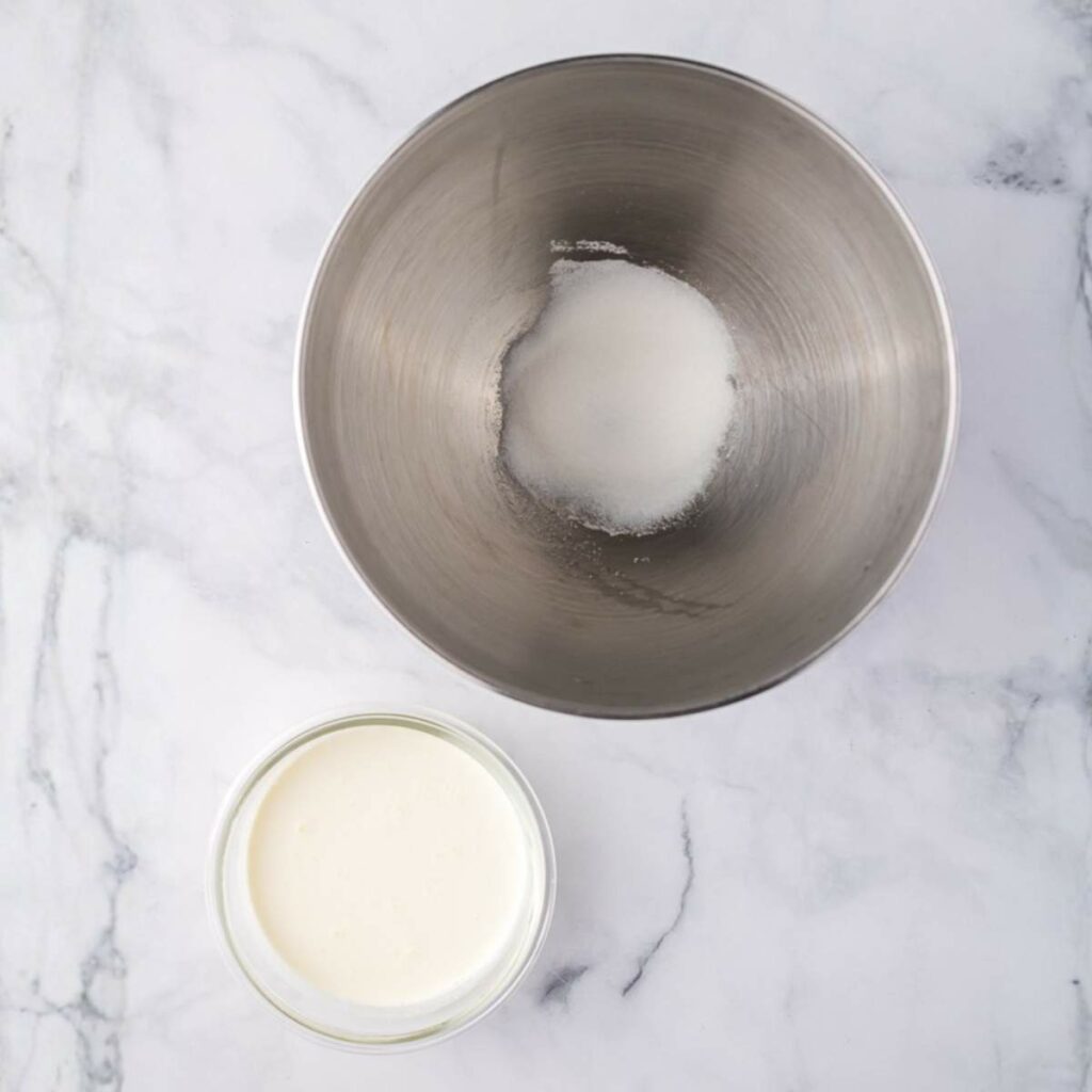 A metal mixing bowl with a small amount of sugar inside, next to a glass bowl filled with cream, on a white marble surface.