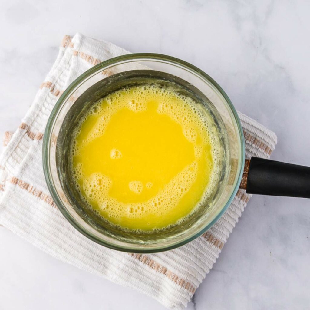 A glass saucepan filled with melted butter sits on a white and beige striped towel over a marble surface.