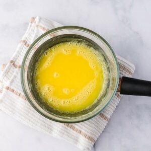 A glass saucepan filled with melted butter sits on a white and beige striped towel over a marble surface.
