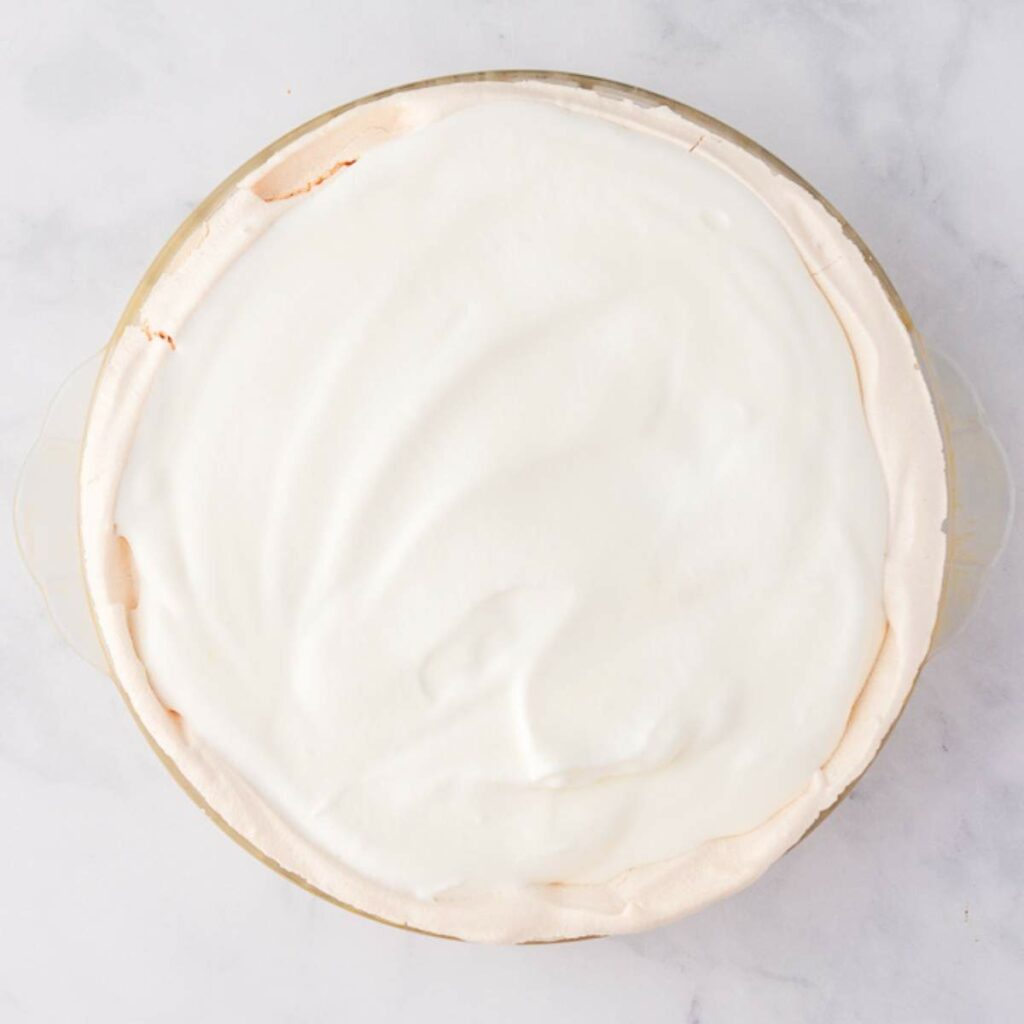 A pie with a smooth, white meringue topping in a glass pie dish, photographed from above on a light marble surface.