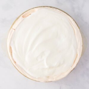 A pie with a smooth, white meringue topping in a glass pie dish, photographed from above on a light marble surface.