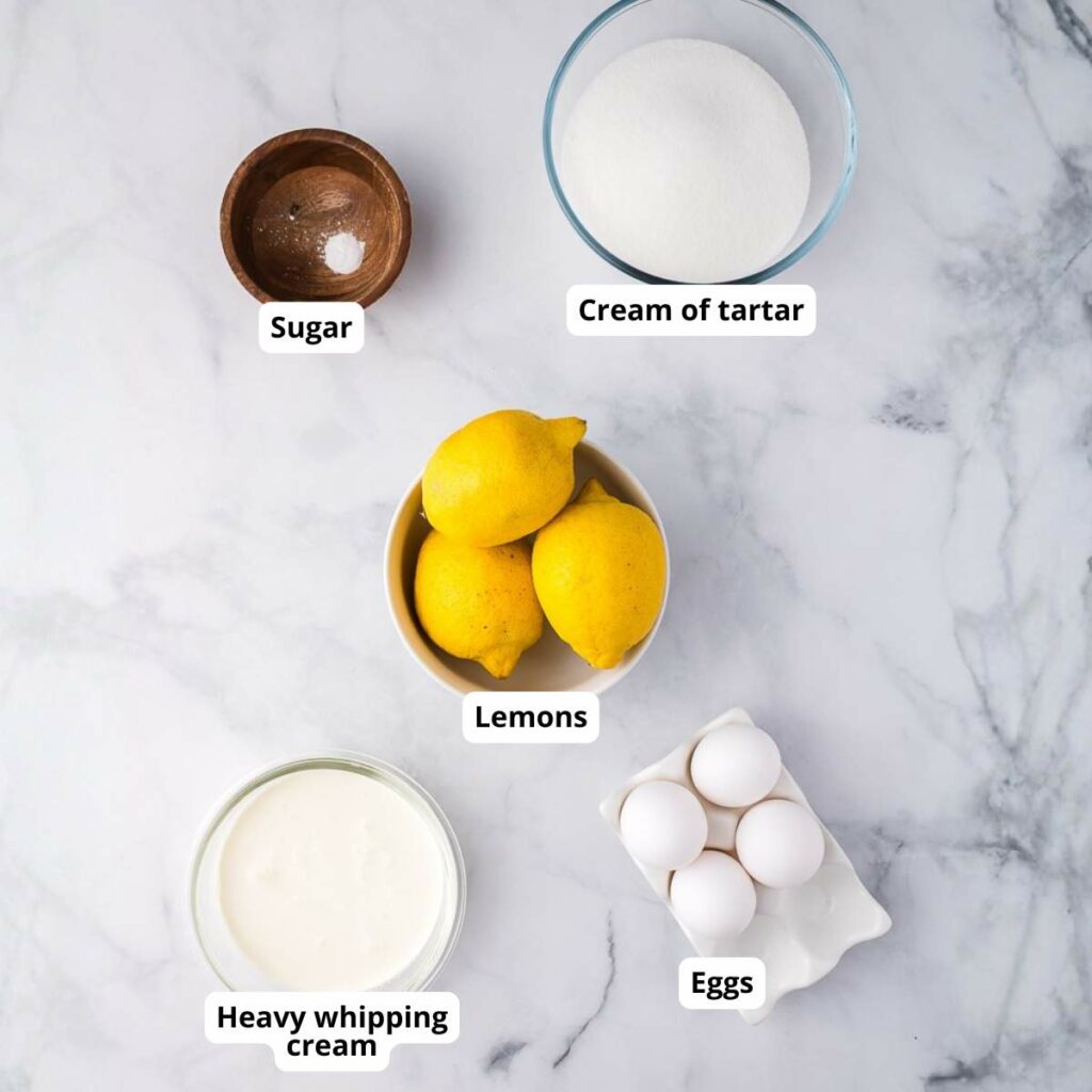 Marble surface with labeled bowls containing sugar, cream of tartar, lemons, heavy whipping cream, and eggs arranged neatly.