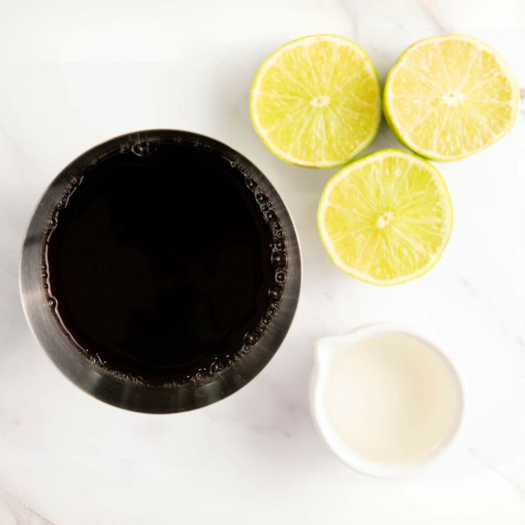 A metal bowl filled with a dark liquid, a small white jug with white liquid, and three halved limes on a white surface.