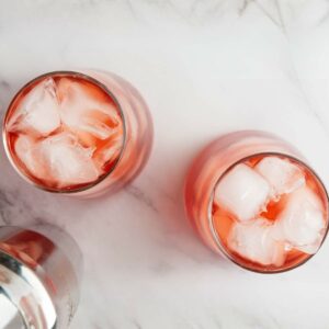 Two glasses filled with ice and a pink beverage are on a marble surface.