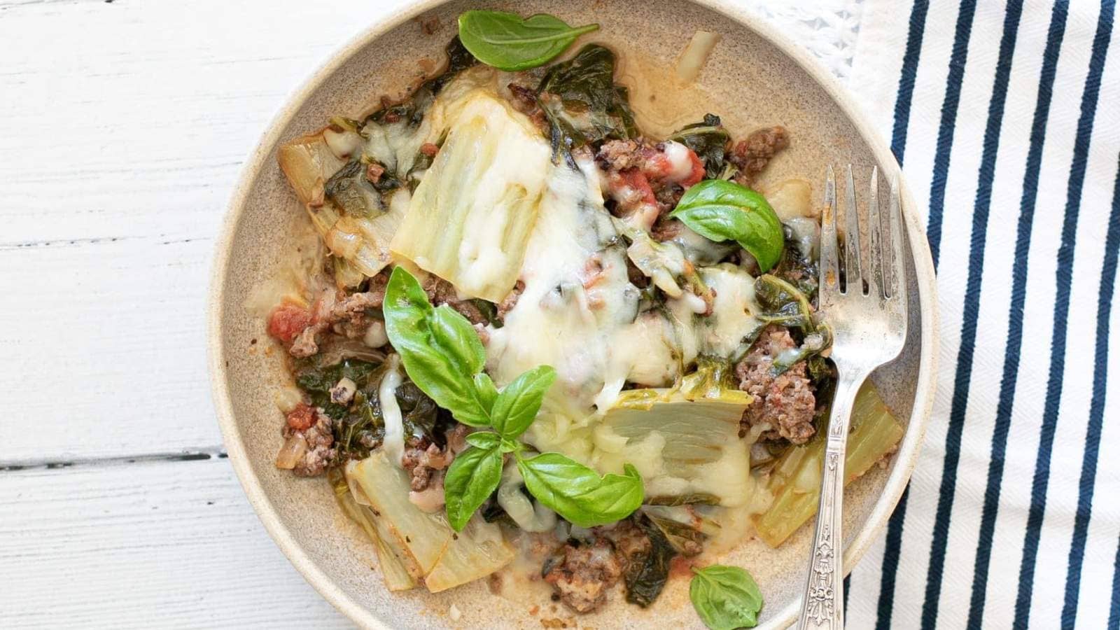 A plate of baked lasagna with ground meat, spinach, melted cheese, tomato sauce, and fresh basil, served with a fork on a light-colored plate.