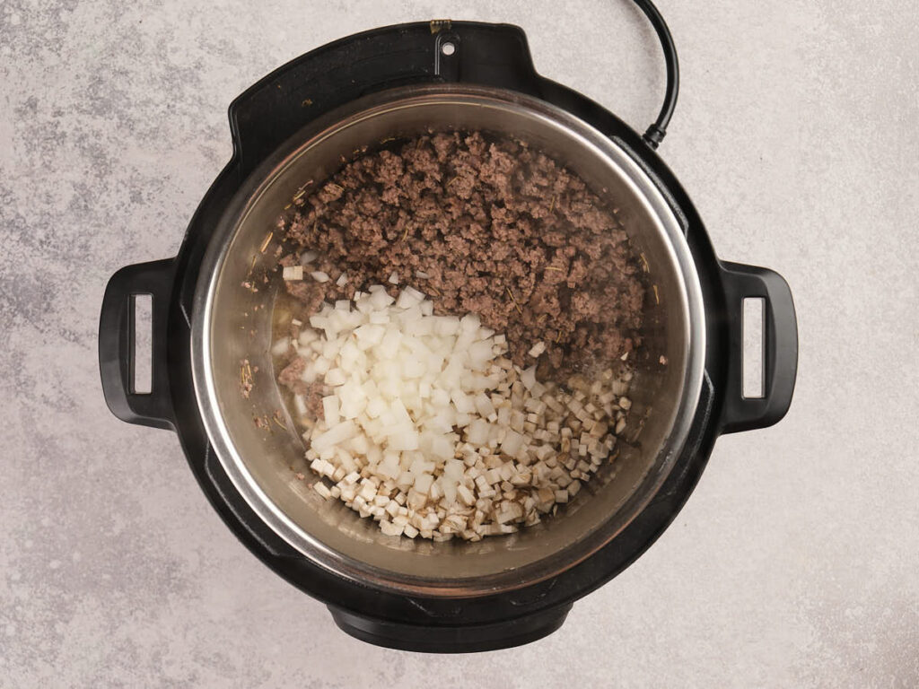 An overhead view of an electric pressure cooker containing cooked ground beef, diced onions, and diced celery on a light countertop.