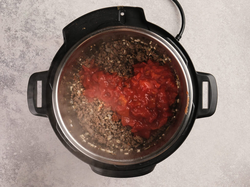 Ground beef, onions, and diced tomatoes are cooking together inside an open electric pressure cooker.