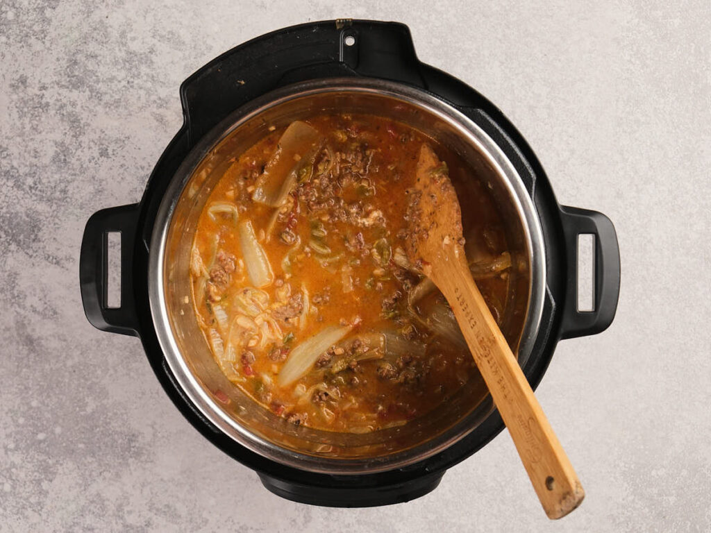 A top-down view of a pot of soup with pasta, vegetables, and ground meat, being stirred with a wooden spoon on a gray surface.