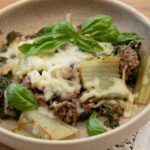 A bowl of baked ground meat, leafy greens, melted cheese, and fresh basil leaves, served on a light brown plate.