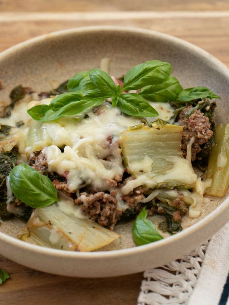 A bowl of baked ground meat, leafy greens, melted cheese, and fresh basil leaves, served on a light brown plate.