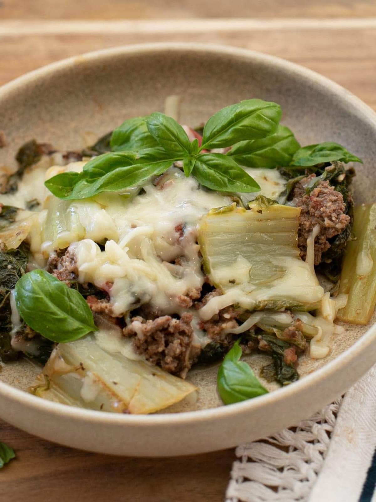 A bowl of baked ground meat, leafy greens, melted cheese, and fresh basil leaves, served on a light brown plate.