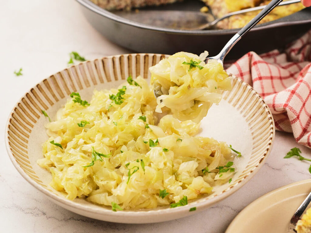 A bowl of cooked cabbage garnished with chopped parsley, with a spoon lifting a portion.