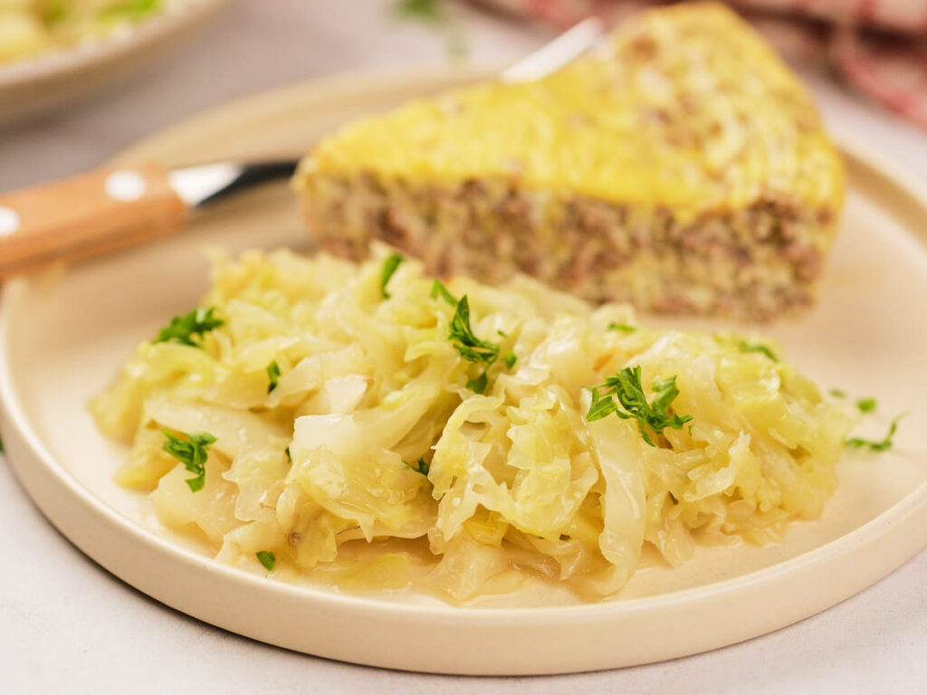A plate with cooked cabbage garnished with herbs, served with a wedge of a baked casserole or meat pie in the background.