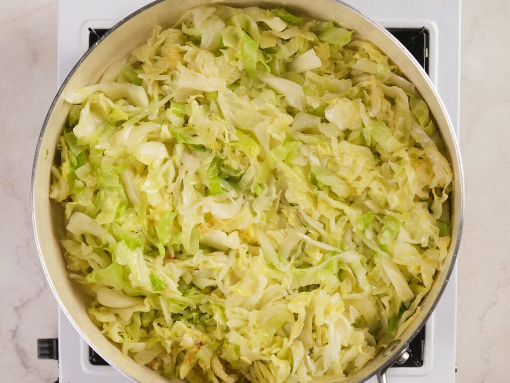 A large pan of cooked, shredded cabbage sits on a stove burner against a light countertop background.