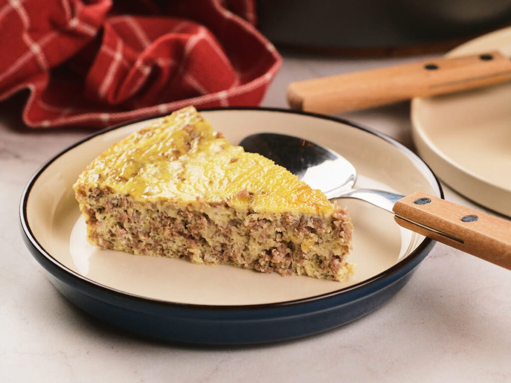 A slice of baked egg and ground meat casserole sits on a plate with a serving utensil beside it. A red checkered cloth and stacked plates are in