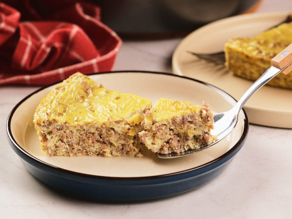 A slice of baked egg and meat casserole is served on a plate, with a fork lifting a bite-sized piece.