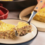 A close-up of a fork holding a bite of a baked casserole with ground meat and egg.