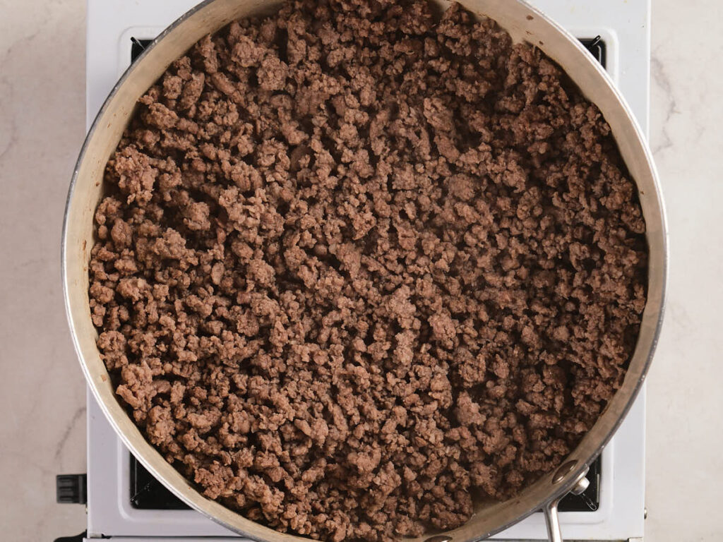 A stainless steel skillet filled with cooked ground beef sits on a white stovetop.