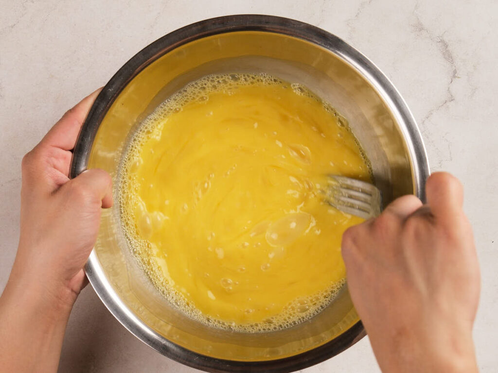 Hands whisking beaten eggs in a stainless steel bowl on a light-colored countertop.