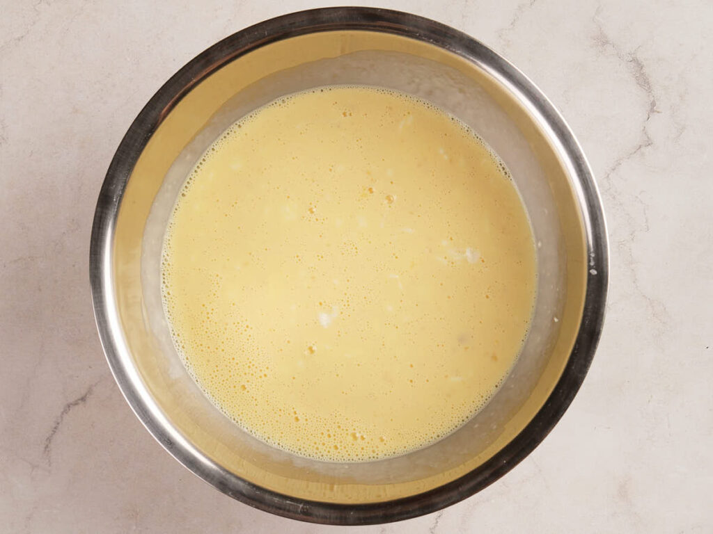 A metal mixing bowl filled with a pale yellow liquid mixture, likely a batter or custard, sits on a light-colored countertop.
