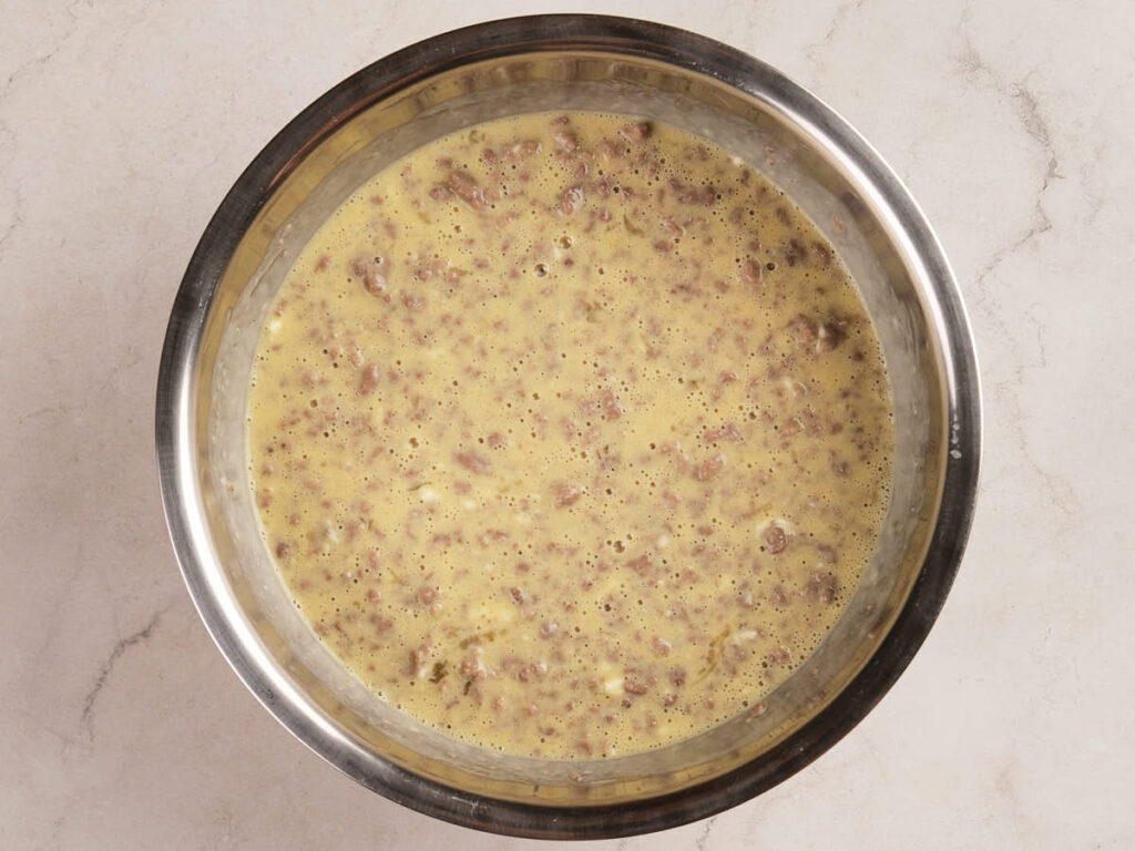 A metal bowl containing a mixture of beaten eggs and browned ground beef on a light-colored countertop.