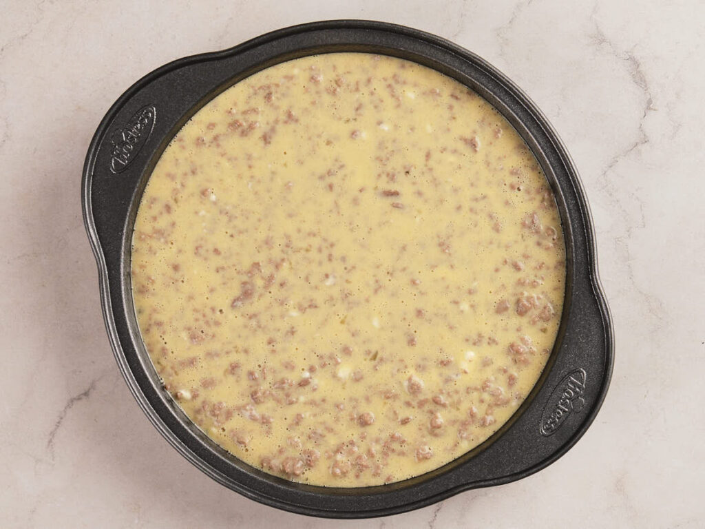 A round black baking pan filled with an uncooked egg and sausage mixture sits on a light-colored countertop.
