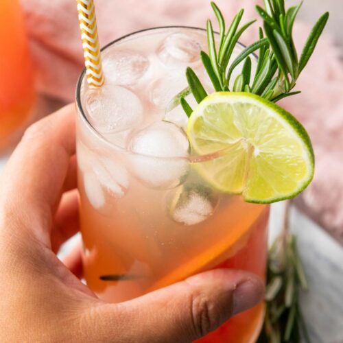 A hand holds a glass of iced beverage garnished with a lime slice and rosemary sprig, with a paper straw in the drink.