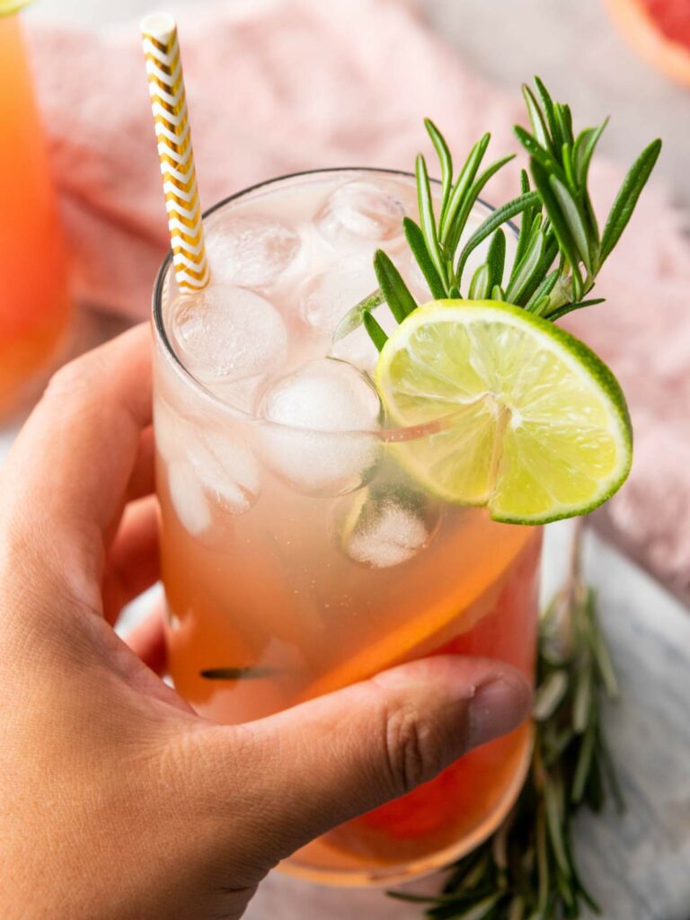 A hand holds a glass of iced beverage garnished with a lime slice and rosemary sprig, with a paper straw in the drink.