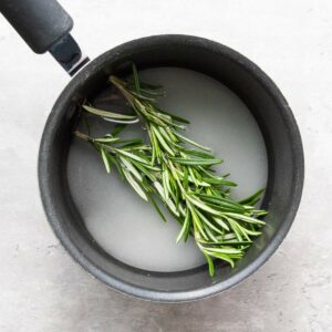 A small saucepan containing water, sugar, and fresh rosemary sprigs on a light-colored surface.