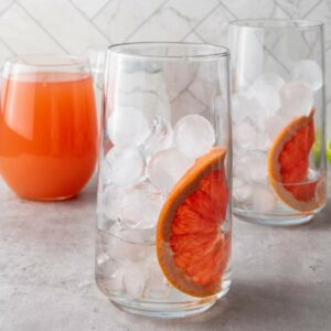A glass filled with ice and a slice of grapefruit, with another similar glass and a glass of orange juice in the background.