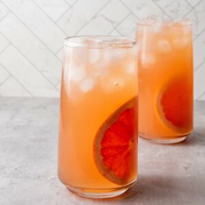 Two tall glasses filled with an orange-pink iced beverage, each garnished with a slice of grapefruit.