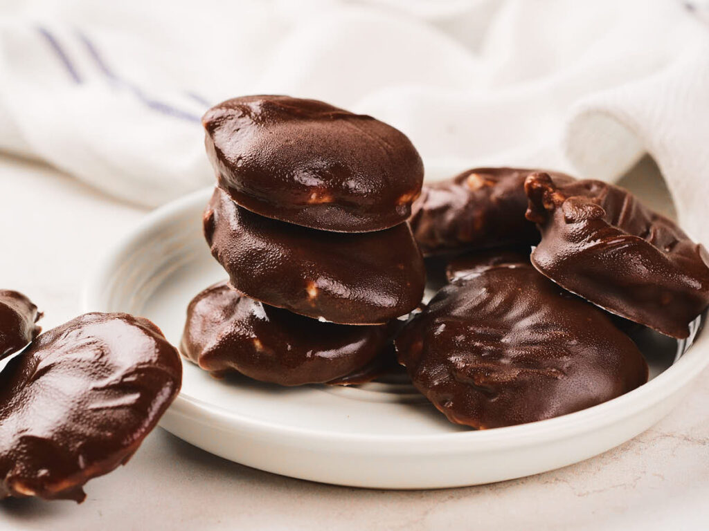 A white plate holds several chocolate-coated pecan pralines, with a white cloth in the background.