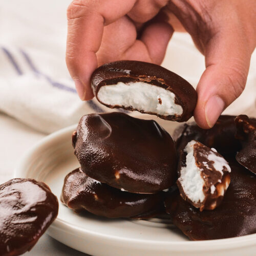 A hand holding a chocolate-covered candy with a white marshmallow center, above a plate stacked with similar candies.