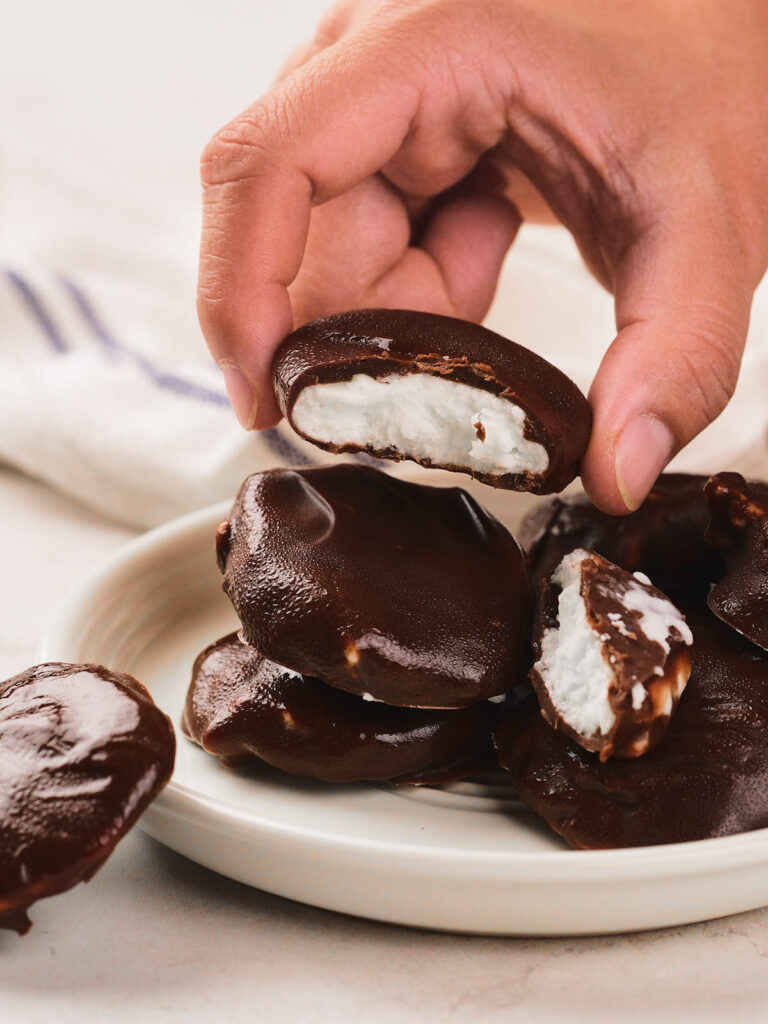 A hand holding a chocolate-covered candy with a white marshmallow center, above a plate stacked with similar candies.