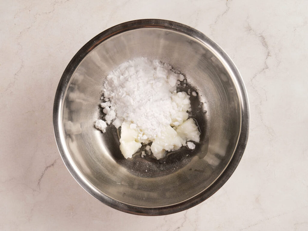 A metal mixing bowl containing white solid chunks and a pile of white powder on a light countertop.
