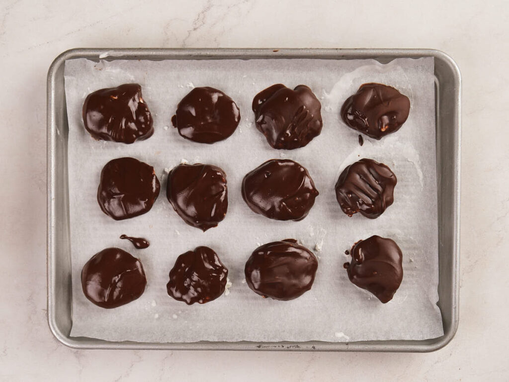 A baking tray lined with parchment paper holds twelve round treats covered in glossy chocolate coating.
