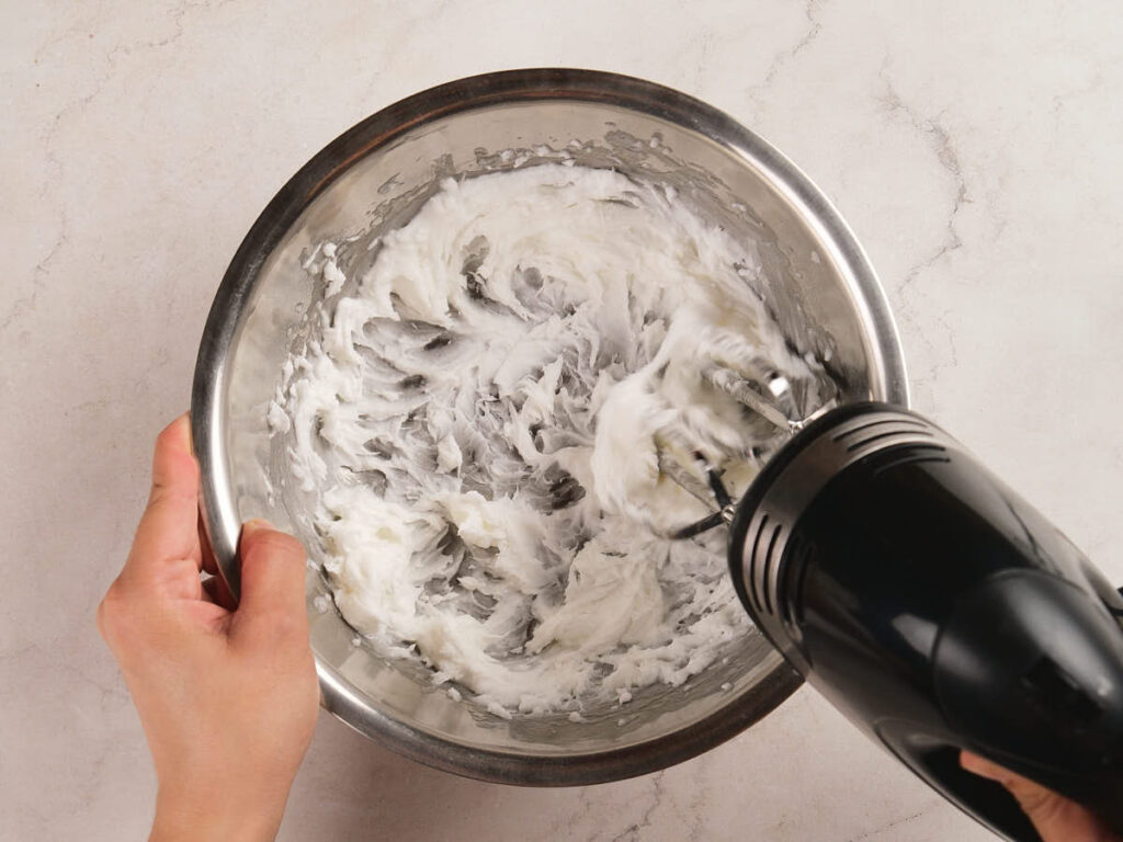 A person uses a hand mixer to blend a white creamy mixture in a stainless steel bowl on a light countertop.