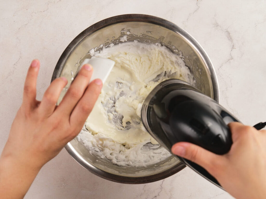 Person mixing white cream in a metal bowl with an electric hand mixer, using a spatula to scrape the sides.