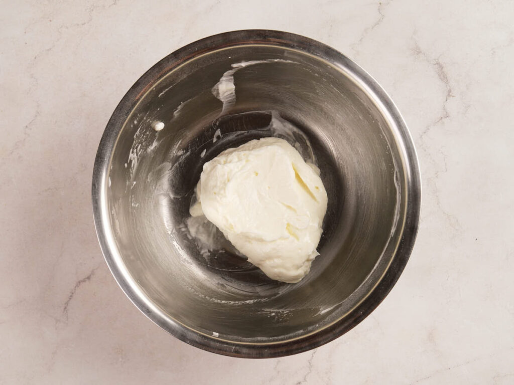 A stainless steel mixing bowl contains a mound of thick, white yogurt on a light marble surface.