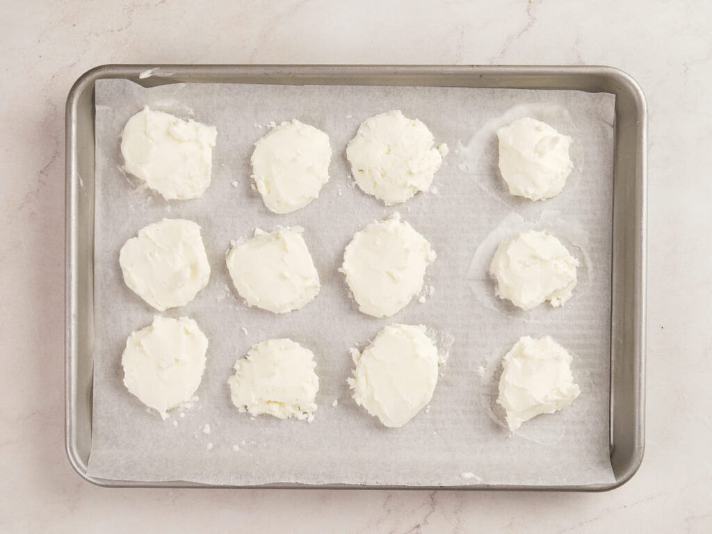 Twelve round portions of white cheese spread are arranged on a parchment-lined baking sheet placed on a light-colored surface.